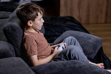 Boy is sitting on sofa at home and playing video games with his joypad.