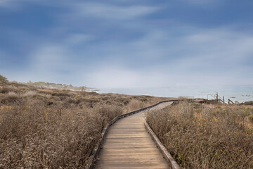 An empty boardwalk leading to the ocean, low brown brush in foreground, , blue wisp clouds and fog...
