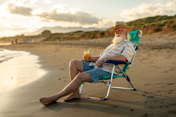 Relaxed senior man with white beard and straw hat sitting in a beach chair by the sea, holding orange juice, enjoying peaceful sunset moments and retirement lifestyle on a calm summer evening.