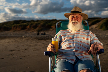 Elderly man with long white beard sitting on beach chair, wearing striped shirt and straw hat, holding glass of orange juice, enjoying calm evening by the sea under soft golden sunlight.