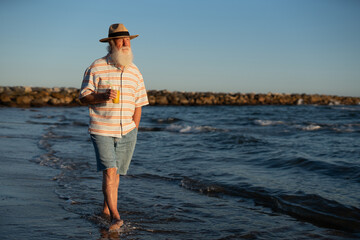 Happy elderly man in a straw hat and striped shirt walking barefoot through shallow sea water,...