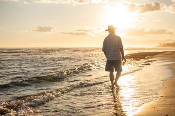 Elderly man in a straw hat walking barefoot along the beach shoreline at golden hour, holding a glass of orange juice and enjoying the peaceful sunset over the calm sea.