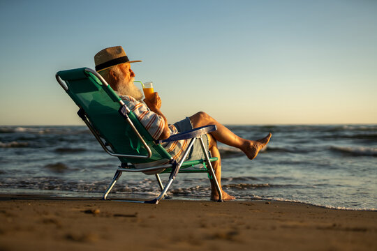 Senior man with white beard sitting in beach chair, sipping orange juice through straw, relaxing by the ocean during sunset, enjoying peaceful moment and listening to sea waves in solitude.