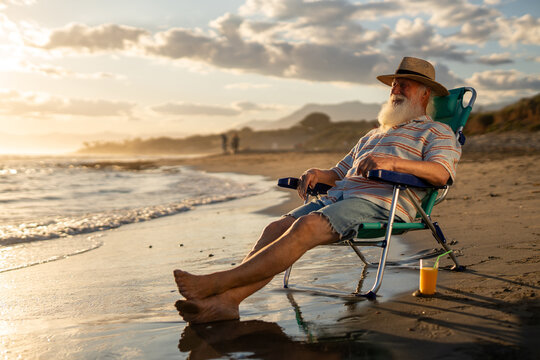 Elderly man in straw hat relaxing on beach chair at golden sunset, eyes closed with peaceful smile, enjoying serene ocean breeze and vacation vibe, with orange juice on sand beside him.