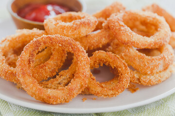 Homemade crispy fried breaded onion rings on a plate with a bowl of ketchup (Selective Focus, Focus on the upper part of the onion ring in the front and the lower part of the one next to it)