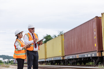 Two engineers wearing safety gear inspect freight containers on a railway track, discussing transport and export logistics. 