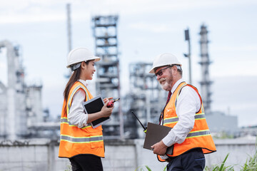 Two engineers wearing safety vests and helmets smile during a discussion at an oil refinery. They...