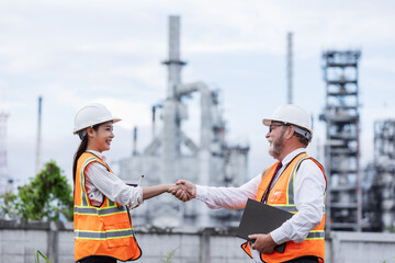 Two engineers shake hands at an oil refinery site, symbolizing successful collaboration or agreement. Both wear safety gear, with refinery structures visible in the background.