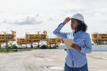 A female engineer wearing a safety helmet holds a tablet while inspecting the construction site of oil rigs. Heavy structures in the background highlight offshore industry development.
