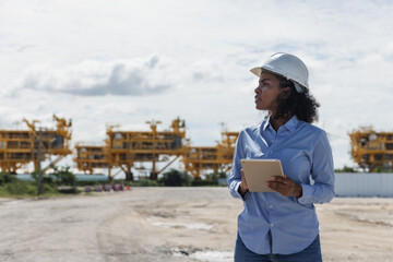 A female engineer wearing a safety helmet holds a tablet while inspecting the construction site of oil rigs. Heavy structures in the background highlight offshore industry development.