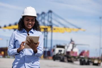 A female engineer in a safety helmet uses a digital tablet while monitoring container logistics at a shipping port. Cranes and trucks in the background show active freight operations.