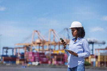 A concerned female engineer at a shipping port checks her phone and tablet, possibly addressing a...