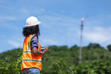 A female environmental engineer in safety gear takes notes outdoors near a forested area with a visible industrial chimney, indicating monitoring or assessment of air pollution impact.