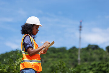 A female environmental engineer in safety gear takes notes outdoors near a forested area with a...