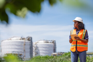 An environmental engineer wearing safety gear uses a tablet while surveying an oil tank facility surrounded by greenery. The image highlights environmental monitoring near industrial infrastructure.