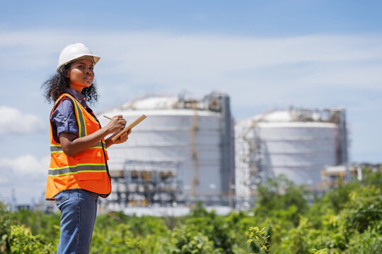 An environmental engineer conducts a site survey near large industrial storage tanks. Wearing safety gear and holding a tablet, she evaluates environmental impact and infrastructure integrity.