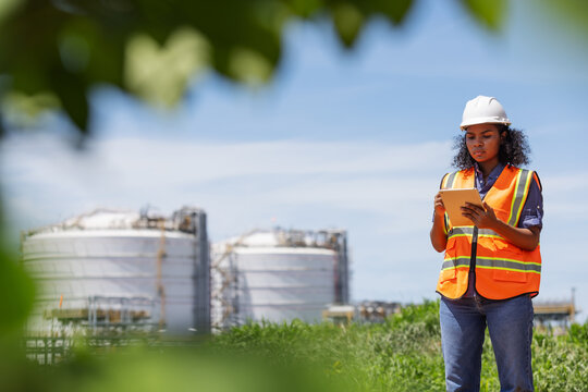 An environmental engineer wearing safety gear uses a tablet while surveying an oil tank facility surrounded by greenery. The image highlights environmental monitoring near industrial infrastructure.