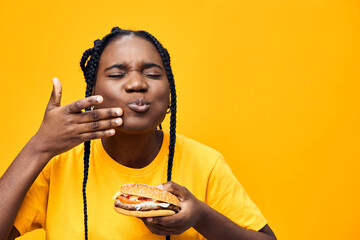 Happy young woman enjoying a delicious burger against a vibrant yellow background, showcasing her joy and love for fast food with playful expressions