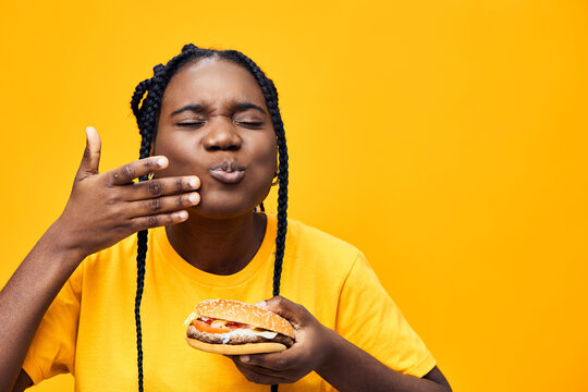 Happy young woman enjoying a delicious burger against a vibrant yellow background, showcasing her joy and love for fast food with playful expressions
