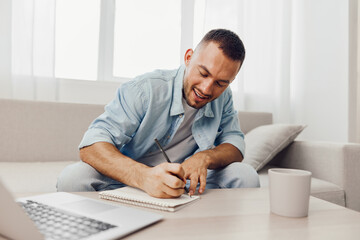Smiling man engaged in creative work at home, using a notebook and laptop, surrounded by a cozy living room environment, reflecting positivity and productivity.