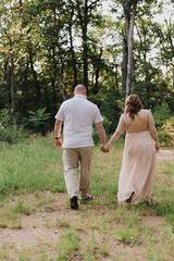 Fototapeta premium Back view of couple walking hand in hand along grassy forest path, woman in long lace-back pink dress and man in white polo with khaki pants under warm afternoon light