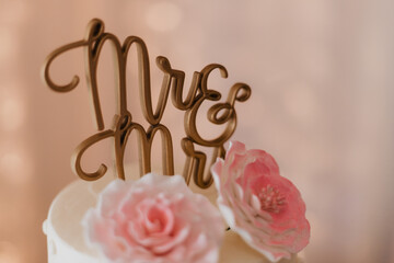 Close-up of white wedding cake topped with gold script “Mrs & Mr” cake topper and pink sugar...