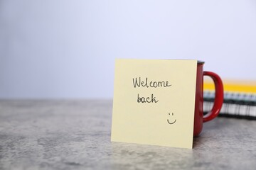 Sticky note with phrase Welcome Back and coffee on grey table, closeup. Space for text