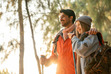 Romantic Couple Hiking in the Forest During Golden Hour. Embracing While Watching Sunset Light Through Trees, Wearing Warm Jackets and Backpacks, Feeling Connected and Adventurous in Nature