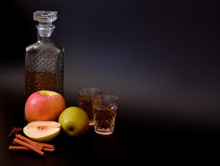 Apple liqueur in a crystal decanter and two glasses on a black background, next to pieces of ripe fruit and cinnamon with anise.
