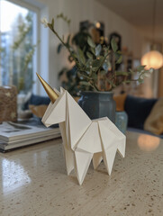 White origami unicorn with gold horn standing on tabletop surface, soft light creates magical feeling of fantasy and wonder, creative paper craft decoration indoors