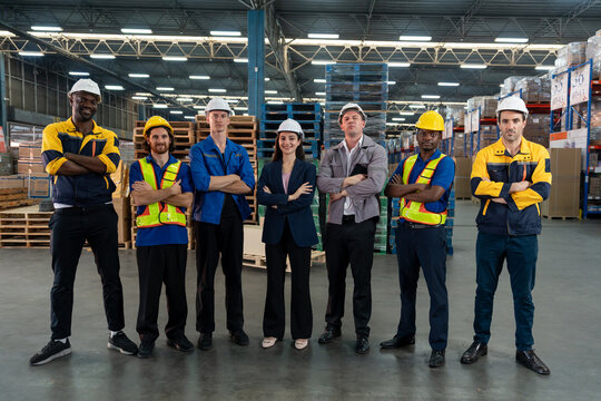 Diverse group of warehouse staff and supervisors standing confidently with arms crossed inside storage facility. Wearing safety uniforms and helmets, representing teamwork, leadership, and cooperation