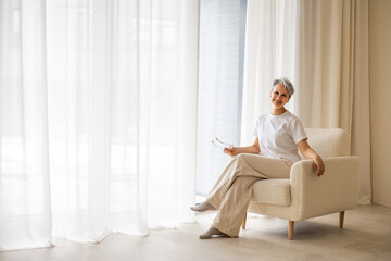Cheerful senior woman in white room sitting on cozy armchair near window holding eyeglasses smiling