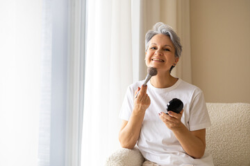 Smiling senior woman applying makeup with brush while sitting in armchair in bright home interior
