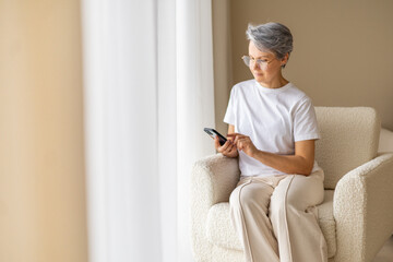 Senior woman using smartphone at home sitting in soft armchair near window in modern bright room