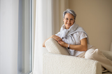 Smiling middle-aged woman with glasses relaxing on a cozy armchair near window at home interior