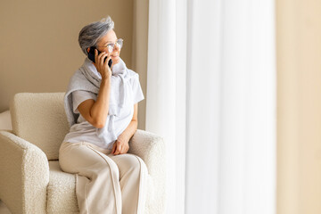 Smiling mature woman talking on smartphone while sitting on cozy armchair by bright window light
