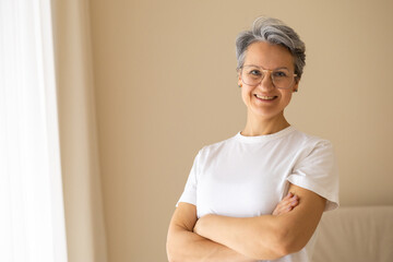 Confident senior woman in white t-shirt smiling with arms crossed, standing in bright modern room