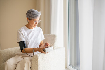 Senior woman checking blood pressure at home using digital monitor while sitting in cozy modern room