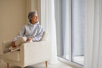 Peaceful mature woman in hoodie sitting in cozy chair looking out large window at home interior