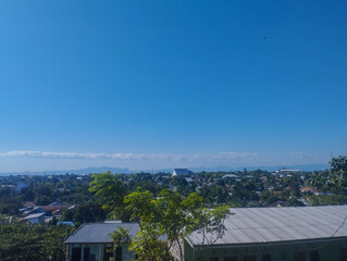 A clear blue sky above a peaceful small town with lush green trees, corrugated metal roofs, and distant mountain horizon