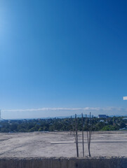 Open construction site with vertical steel rebars in the foreground and a panoramic view of a tropical town under a clear blue sky with distant mountains