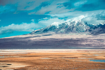 Golden Valley Wetland in Taxkorgan: Serene marshland reflecting snow-capped Pamir Mountains under clear blue sky with drifting clouds. Xinjiang, China natural landscape.