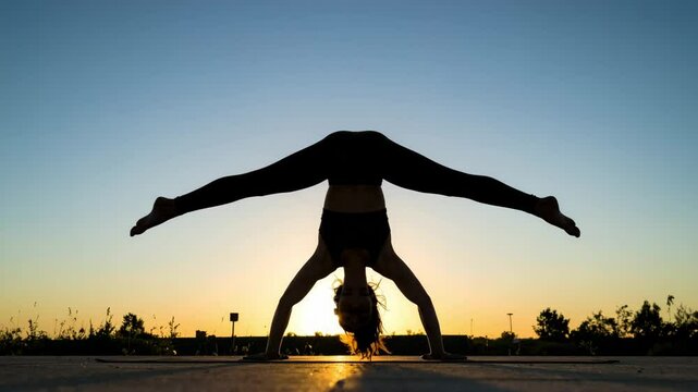 A person performs a handstand split outdoors at sunset, silhouetted against the glowing sky.