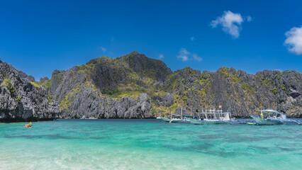 Bangka boats are anchored in the aquamarine ocean. There is green tropical vegetation on the steep coastal cliffs. Blue sky, clouds. Philippines. Palawan.