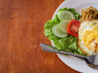 A dish of Indonesian noodles topped with a perfectly fried egg. Served on a clean white plate, this dish is complemented with fresh cucumber, lettuce, and tomato.