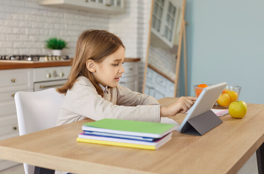 Small cute child engaged in online study activity on tablet, learning, playing, watching video lesson, sitting at table in modern kitchen setting, distance learning courses, webinar, digital resource - Powered by Adobe