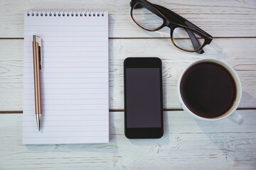 Flat lay is showing spiral notebook with pen on white table with smartphone, eyeglasses, coffee mug