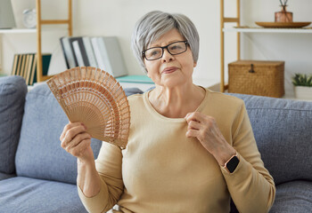 Elderly woman with short gray hair sitting on couch in warm living room and waving wooden hand fan....