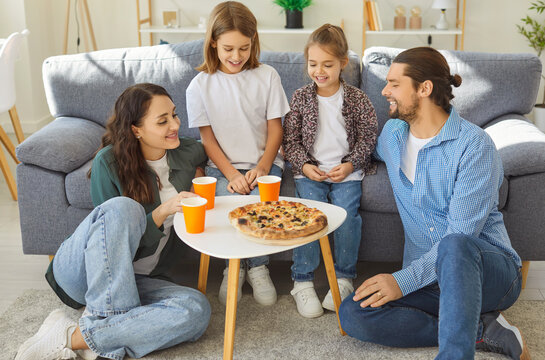 Happy smiling family sitting around coffee table, sharing hot pizza and drinks during weekend lunch. Cheerful parents and children enjoying tasty meal together while spending quality time at home.