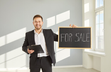 Smiling real estate agent standing in sunny empty apartment, holding clipboard and blackboard with...